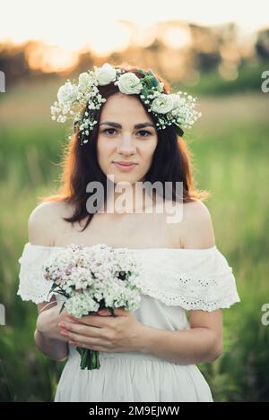 Portrait eines attraktiven Mädchens in einem weißen Kleid mit Blumenstrauß und einem Kranz im Sommer auf dem Feld Stockfoto