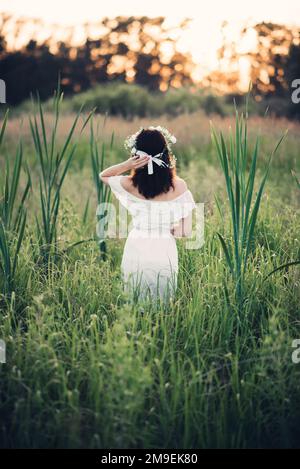 Ein Mädchen in einem weißen Kleid mit Blumenkranz steht im Sommer auf dem Feld. Das Konzept von Einheit, Liebe und Fürsorge für die Natur Stockfoto