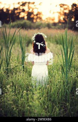 Ein Mädchen in einem weißen Kleid mit Blumenkranz steht im Sommer bei Sonnenuntergang auf dem Feld Stockfoto