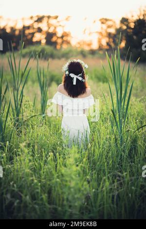 Ein Mädchen in einem weißen Kleid mit Blumenkranz steht im Sommer auf dem Feld. Das Konzept von Einheit, Liebe und Fürsorge für die Natur Stockfoto