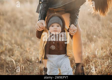 mama hält ihren kleinen Jungen an den Händen für einen Spaziergang im Herbst in der Natur. Die ersten Schritte des Babys Stockfoto