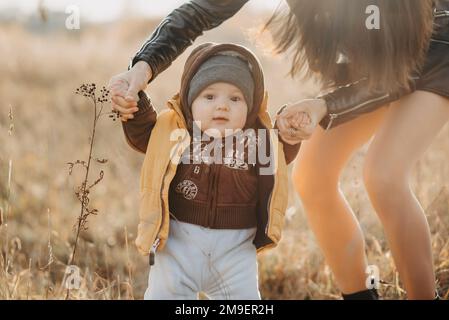 mama hält ihren kleinen Jungen an den Händen für einen Spaziergang im Herbst in der Natur. Die ersten Schritte des Babys Stockfoto