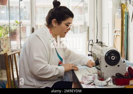 Profilansicht einer jungen Kaukasierin, Freiberuflerin argentinischer ethnischer Herkunft, sitzend, konzentriert arbeiten, mit ihrer Nähmaschine nähen Stockfoto
