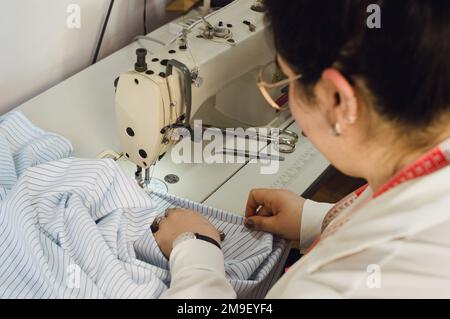 Rückansicht einer jungen, kaukasischen Näherin, die in ihrer Heimwerkstatt arbeitet, ein Tuch mit einer Nähmaschine näht, Textilkonzept, Kopierraum. Stockfoto