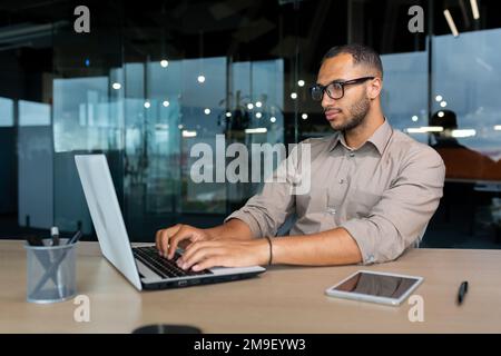 Seriöser und konzentrierter hispanischer Programmierer, der im Büro arbeitet, Mann im Hemd, der Codes für Software mit einem Laptop schreibt. Stockfoto