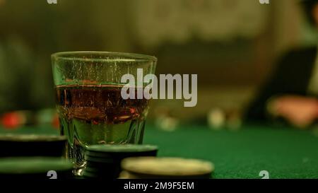 Ein Mann schüttet Whiskey in ein Glas, das auf einem Pokertisch mit grünem Filz steht. Um das Glas herum sind Stapel von Pokerchips, die auf einen Gam hindeuten Stockfoto