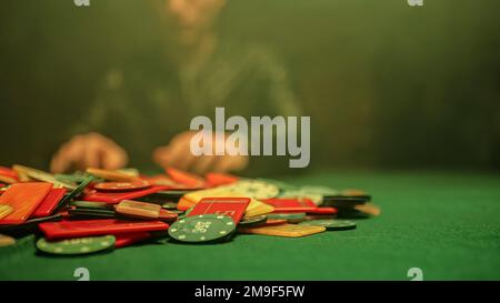 Eine chaotische Szene eines Pokerspiels mit Chips, die willkürlich über den Tisch verteilt sind. Verschwommener Hintergrund eines Pokerspielers, der am Tisch sitzt. Stockfoto