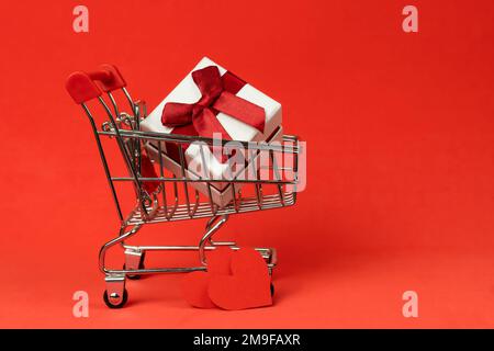 Warenkorb mit weißer Geschenkbox auf rotem Hintergrund, Kopierbereich. Konzept von Geschenken, Liebe, Feiertagen, Valentinstag Stockfoto