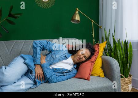 Hispanische Frau krank, die auf dem Sofa im Wohnzimmer liegt, Frau hat starke Magenschmerzen, verärgert, die Hände an den Bauch zu halten. Stockfoto