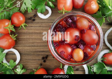 Ernte, Konserven, Salzen oder Beizen roter Tomaten. Tomaten in einem Glasgefäß mit Gurken, Zwiebeln, Paprika und Kräutern auf einem alten rustikalen Stockfoto