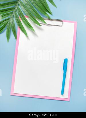 Minimalistisches Konzept des modernen Sommerlayouts von Schreibwaren. Weißer, leerer Rohling und Stift, auf einem rosa Klemmbrett, auf blauem Hintergrund, in der Nähe eines Palmenblattes. Pastellfarben Stockfoto