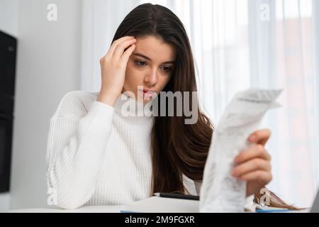 Verärgerte Frau sieht sich nach dem Einkaufen die ausgegebene Summe auf der Rechnung an. Ein junger Freiberufler sitzt neben Notebook und prüft die Liste der Einkäufe zu Hause Stockfoto