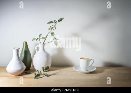 Blasses Stillleben mit drei Vasen in Weiß und Grün, einem Salbeiblatt-Zweig mit Schatten und einer Kaffeetasse, einem Holztisch an einer grauen Wand, machen Sie eine Pause w Stockfoto