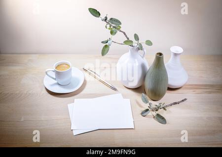 Drei Vasen, Salbeiblatt-Zweig und Kaffeetasse, Modell weißer Grußkarten auf einem Holztisch oder Schreibtisch an einer hellbeigen Wand, ruhiges Home Office Stockfoto