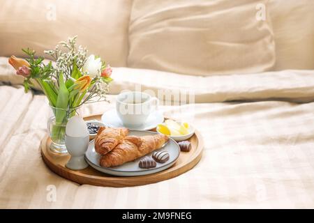 Romantisches Frühstück im Bett mit Blumen, Croissant, Kaffeetasse und Herzen aus Schokolade auf beiger Bettwäsche für Valentinstag oder Muttertag, Kopierbereich, Stockfoto