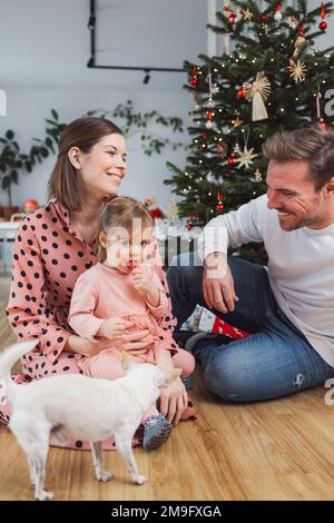 Fröhliche, lächelnde Familie mit einem kleinen Mädchen und einem Hund, die am Weihnachtstag Spaß haben Stockfoto