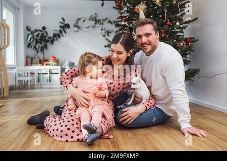 Junge Familie, die auf dem Boden vor dem Weihnachtsbaum sitzt Stockfoto