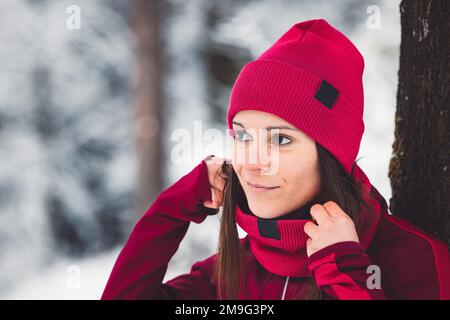 Wunderschöne Brünette mit passendem roten Hut und Schal, während Sie im kalten, verschneiten Wald spazieren Stockfoto