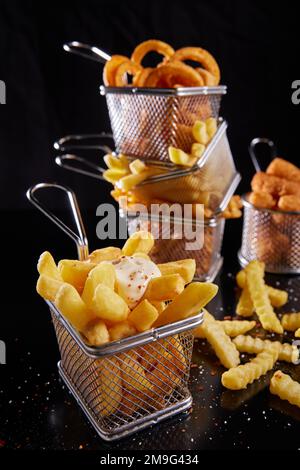 Von oben: Leckere knusprige, salzige Pommes Frites mit Sauce, serviert in einem Metallgitterkorb in der Nähe von Zwiebelringen und Nuggets auf schwarzem Hintergrund Stockfoto