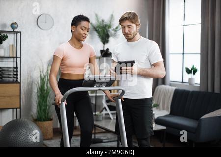 Fitness Kaukasier-Trainer mit Tablet, um eine Online-Lektion für afroamerikanische Frauen zu zeigen, die auf einem Laufband in einem modernen Zuhause trainieren. Zwei sportliche Personen, die sich an Ferntraining beteiligen. Stockfoto