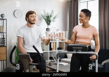 Porträt eines jungen Paares, das auf einem Heimtrainer und einem Laufband Cardio-Training macht und Wasserflaschen knallt, zum Glück freut es sich, das Training zu beenden. Attraktive Frauen und Männer trainieren auf Fitness. Stockfoto
