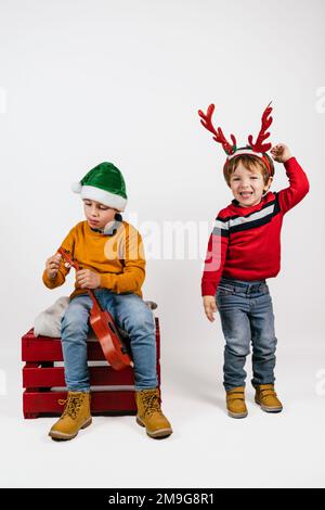 Blonde, weiße, lächelnde Kinder in weihnachtshüten, die auf weißem weihnachtshintergrund spielen Stockfoto