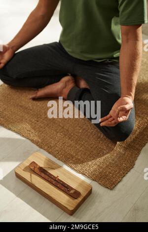 Blick aus einem großen Winkel auf einen jungen Mann, der in Lotusposition auf dem Boden sitzt, mit einem Aromastift duftet und Yoga macht Stockfoto