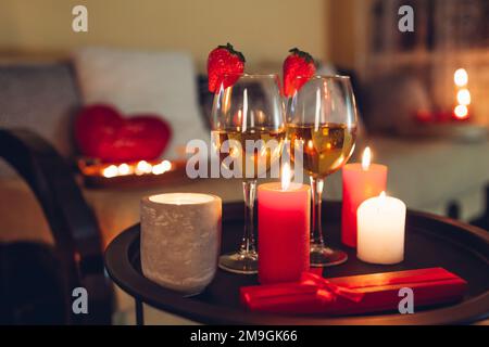 Valentinstag-Feier zu Hause mit Champagnerweingläsern mit Erdbeeren umgeben von Kerzen. Rote Geschenkbox Stockfoto