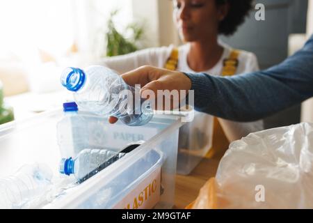 Gekürzte Hand eines birassischen Mannes, der Plastikabfälle in Recyclingbehältern sortiert, mit Freundin zu Hause Stockfoto