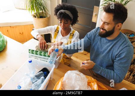 Aus dem großen Blickwinkel sehen Sie ein birassisches junges Paar, das Papier und Plastikmüll in Mülltonnen auf dem Tisch sortiert Stockfoto