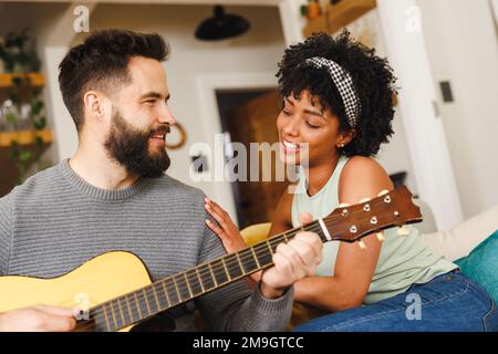 Glücklicher birassistischer junger Mann, der Gitarre spielt, während er mit seiner Freundin auf dem Sofa im Wohnzimmer sitzt Stockfoto