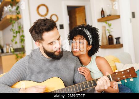 Lächelnde, birassische junge Frau mit Afro-Haaren, die ihren Freund umarmt, singt und Gitarre auf dem Sofa spielt Stockfoto