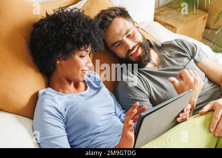 Aus einem großen Blickwinkel ein lächelndes, birassistisches junges Paar, das ein digitales Tablet benutzt, während es zu Hause im Bett liegt Stockfoto