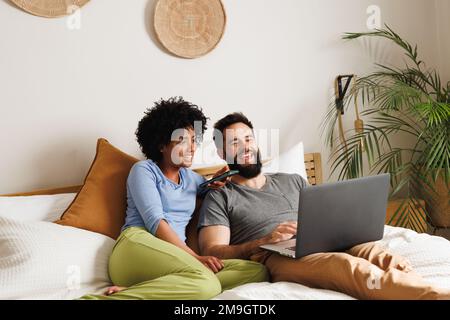 Fröhliches, birassisches junges Paar, das sich über einen Laptop bewegt, während es sich auf dem Bett an der Wand entspannt Stockfoto
