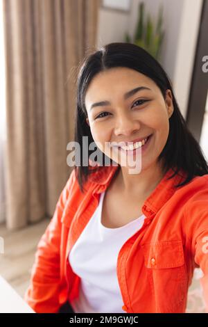 Bild einer lächelnden, birassischen Frau mit dunklem Haar und orangefarbenem Hemd zu Hause Stockfoto