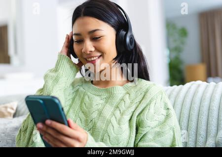 Bild einer glücklichen birassischen Frau mit Kopfhörern, die ihr Smartphone zu Hause auf dem Sofa benutzt, Kopierraum Stockfoto