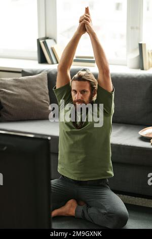 Junger Mann mit Bart, der Yoga-Übungen macht, während er im Wohnzimmer vor dem Fernseher sitzt Stockfoto