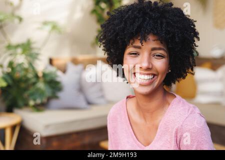 Nahaufnahme einer lächelnden, birassischen jungen Frau mit Afrohaar im Wohnzimmer Stockfoto