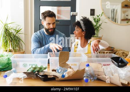 Birassisches junges Paar, das Papier, Kunststoffe und Gläser in Mülltonnen auf dem Tisch diskutiert und sortiert Stockfoto