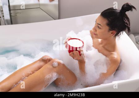 Bild einer birassischen Frau, die sich mit einer Tasse Tee in der Badewanne im Badezimmer entspannt Stockfoto