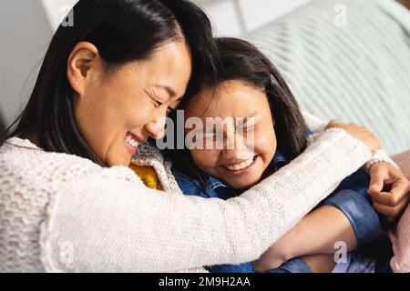 Glückliche asiatische Mutter und Tochter, die auf dem Sofa im Wohnzimmer sitzen und sich umarmen Stockfoto