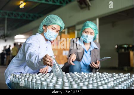 Asiatische Kontrolleurin mit Assistenzangestelltem in sterilen Anzügen, die auf Lagerprodukte der Verpackung von Fruchtgetränken in Lager A prüft Stockfoto