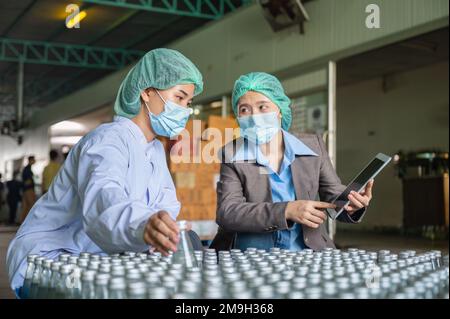 Asiatische Kontrolleurin mit Assistenzangestelltem in sterilen Anzügen, die auf Lagerprodukte der Verpackung von Fruchtgetränken in Lager A prüft Stockfoto