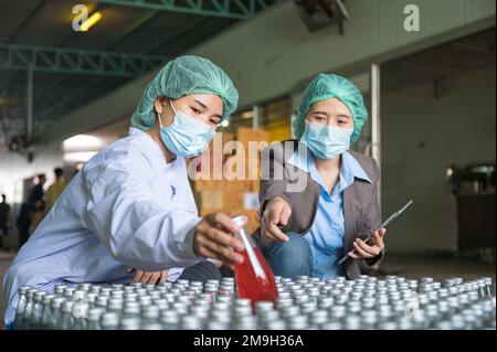 Asiatische Kontrolleurin mit Assistenzangestelltem in sterilen Anzügen, die auf Lagerprodukte der Verpackung von Fruchtgetränken in Lager A prüft Stockfoto