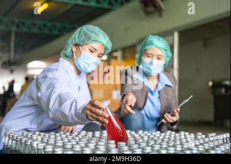 Asiatische Kontrolleurin mit Assistenzangestelltem in sterilen Anzügen, die auf Lagerprodukte der Verpackung von Fruchtgetränken in Lager A prüft Stockfoto