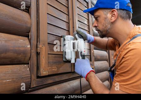 Reparaturarbeiter, der das Holzhaus mit einem Schleifgerät renoviert, bevor er den Fleck zum Wetterschutz aufträgt. Stockfoto