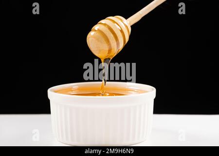 Honig in einer weißen Keramikschüssel mit einem Holzlöffel auf schwarzem Hintergrund. Stockfoto
