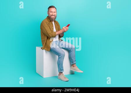 Foto eines positiven erfolgreichen Mannes Tragen trendige Kleidung Sitzen Podium Verwenden Sie moderne Gerätetechnologie isoliert auf cyanfarbenem Hintergrund Stockfoto