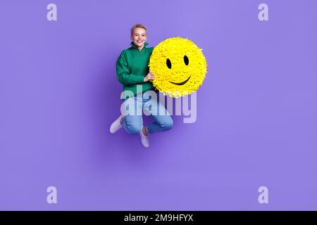 Langes Foto einer positiven, fröhlichen Frau trägt einen grünen Pullover, der hoch springt und einen Smiley Pinata hält. Ein isolierter violetter Hintergrund Stockfoto