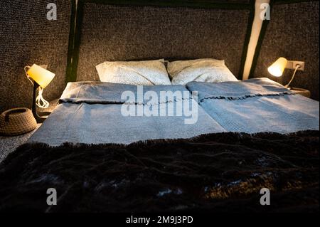 Doppelbett auf Teppichboden im Schlafzimmer der Holzkabine. Pyramidenförmiger Bungalow. Kleines Zimmer, abgewinkelte Wände, Fensterbeleuchtung, Bett, Nachtlampe und Knitte Stockfoto